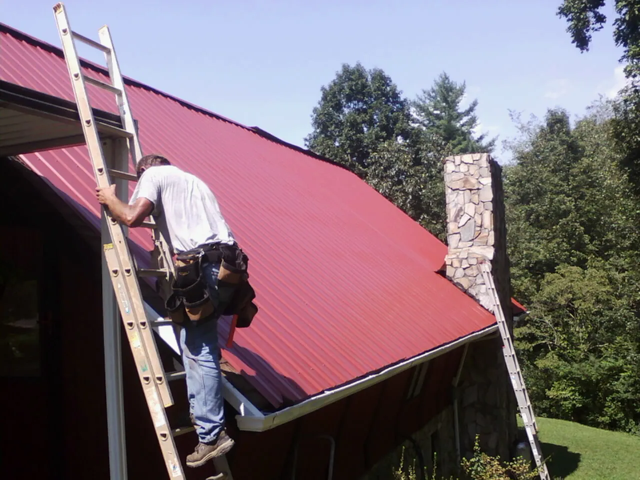 Roofer on a ladder beside a red metal roof and stone chimney — Soffit & Fascia Repair by Roofing Brothers in Berne